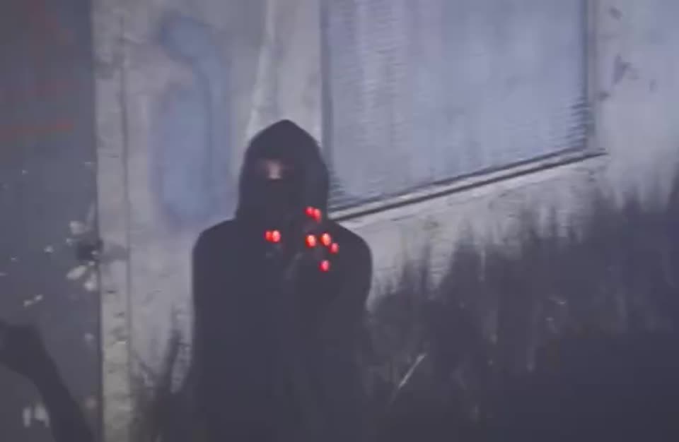 Hooded, masked figure with red LED lights on the mask against a grey concrete wall
